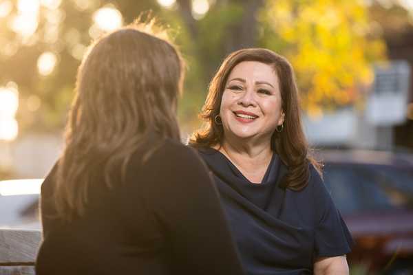 Sandra Armenta smiles while speaking with another person seated across from her on an outdoor bench, with warm evening sunlight filtering through trees in the background.