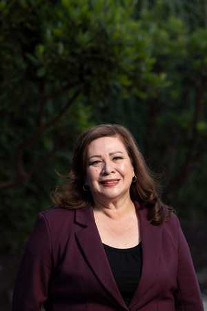 Portrait of Sandra Armenta smiling at the camera while wearing a burgundy blazer over a black top, standing outdoors with a softly blurred background of green trees.