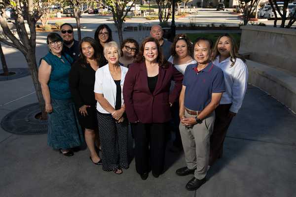 Sandra Armenta stands with a group of community members outdoors for a group portrait in a plaza, with trees, walkways, and campus buildings in the background.