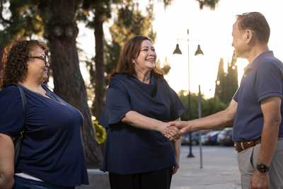 Sandra Armenta smiles as she shakes hands with a community member outdoors while another attendee stands nearby, with trees, parked cars, and evening light in the background.