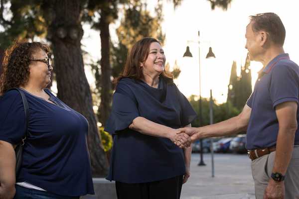 Sandra Armenta smiles as she shakes hands with a community member outdoors while another attendee stands nearby, with trees, parked cars, and evening light in the background.